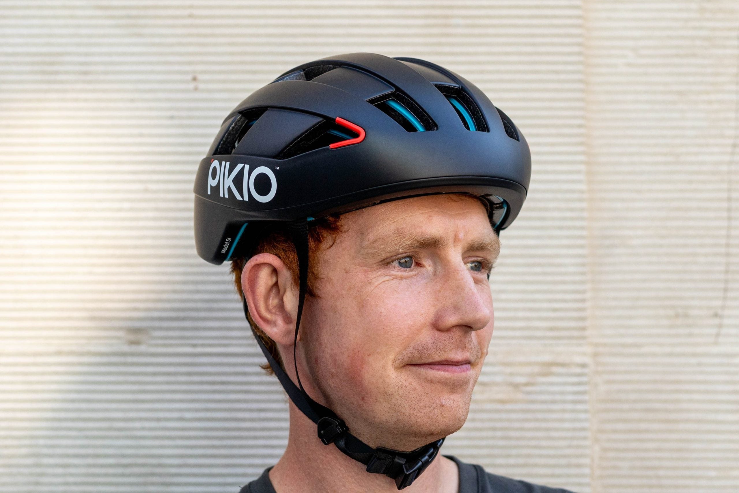 A man wearing a black PIKIO Si OBLIK bicycle helmet, photographed in profile against a light beige textured wall.