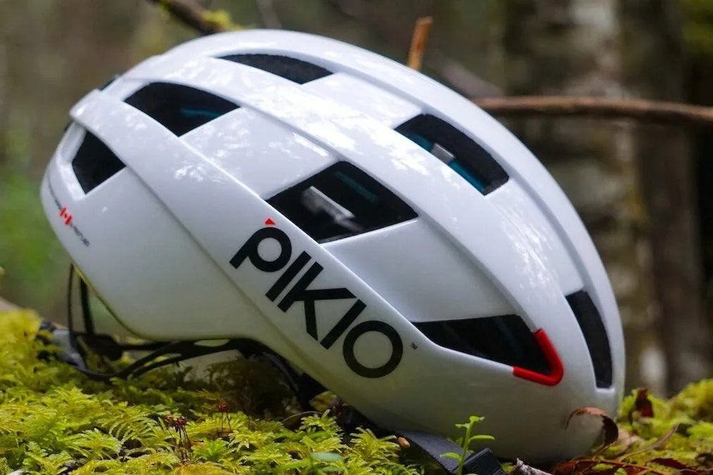 A white PIKIO Si OBLIK bicycle helmet with black and red accents, resting on a bed of green moss in a forest setting.