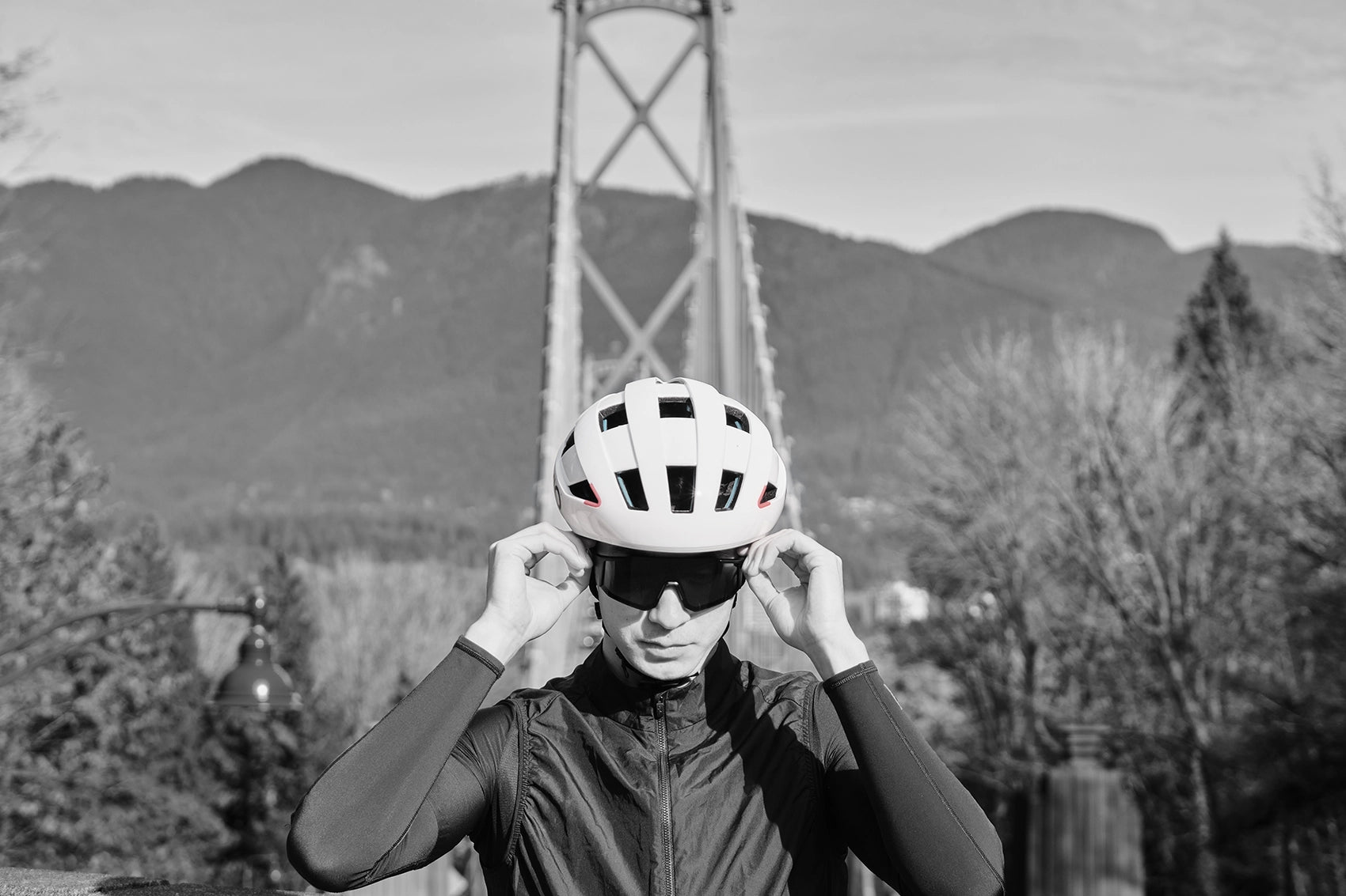 Black and white photo of a man putting on sunglasses while wearing a PIKIO Model Si Helmet in front of a bridge