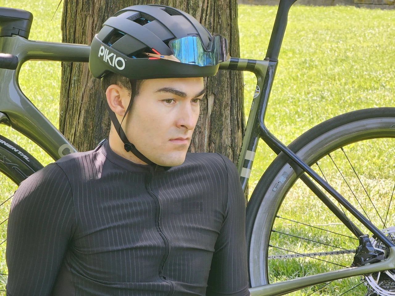Cyclist wearing a PIKIO Si helmet leaning against a tree with a bicycle.