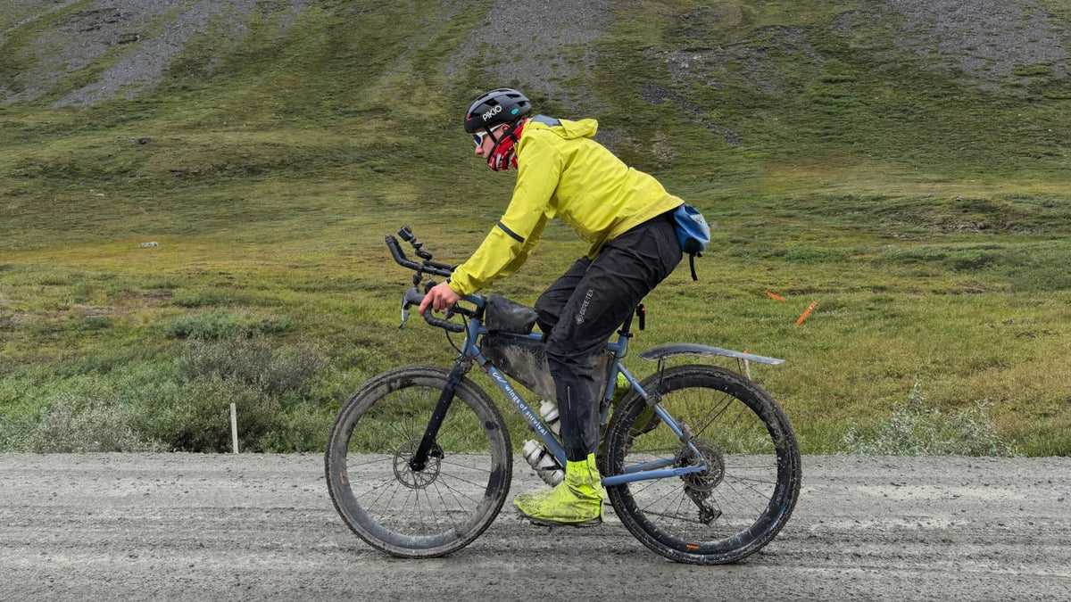 A cyclist wearing a PIKIO Si OBLIK helmet, abright yellow jacket, black helmet, and black pants rides a blue gravel bike on a dirt road with a grassy hill in the background.